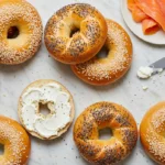 Freshly made bagels with sesame, poppy seeds, and plain toppings styled with cream cheese and smoked salmon on a marble surface.